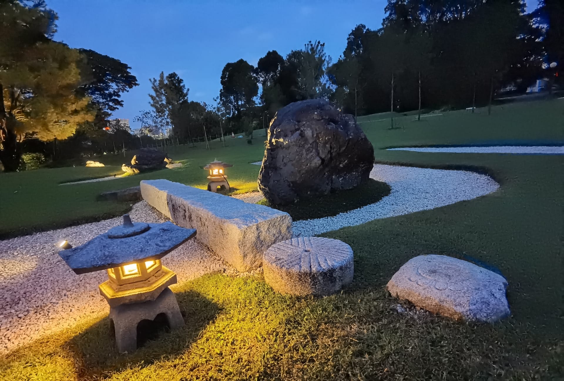 Zen garden with stone lanterns, boulders, and a winding white gravel path at dusk.